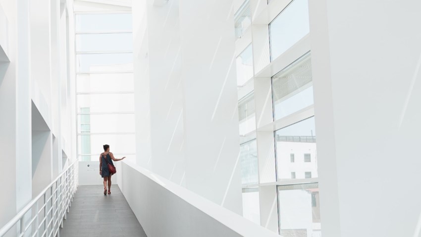 Person walking through a bright modern white hallway with tall glass windows.